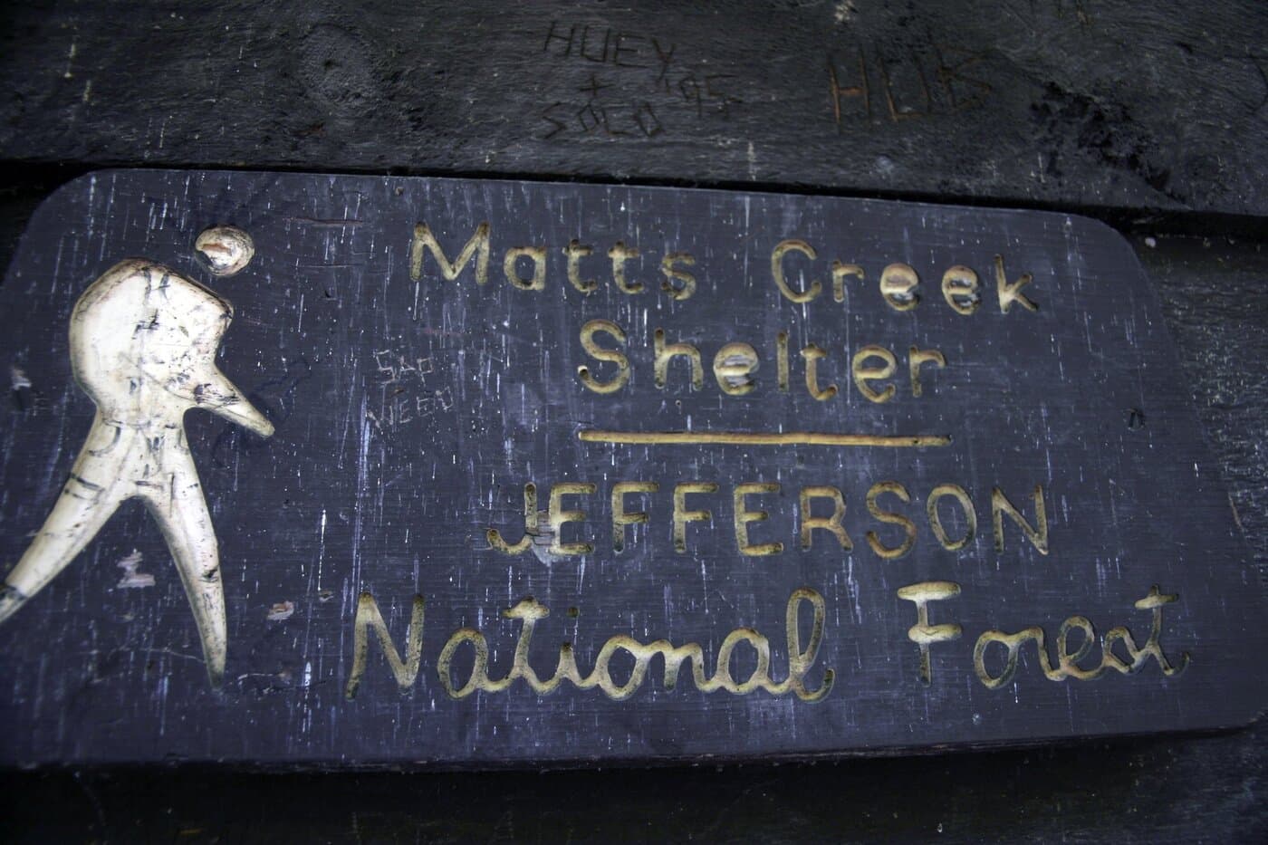 Carved wooden sign reading Matts Creek Shelter, Jefferson National Forest, with a hiker figure, mounted on dark boards scratched with initials.